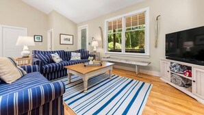A living room with a sofa set, a coffee table, a TV, and a window with a view of greenery. - Union Pier Hidden Gem | 3 Blocks to Beach | Screened Porch & Fire Pit (Union Pier)