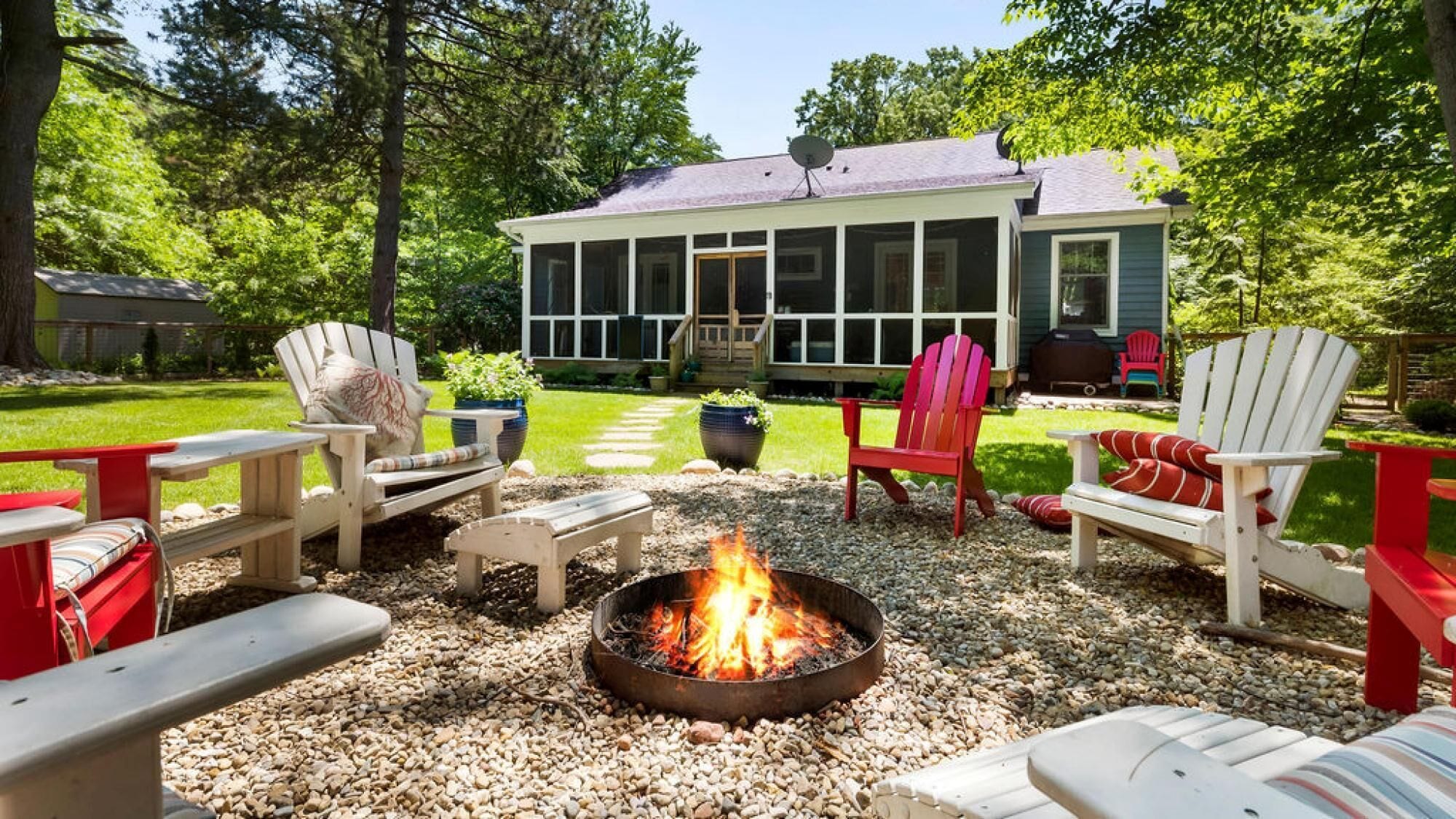 Un patio trasero con un hoyo de fuego, sillas Adirondack y una casa con porche mosquitero.