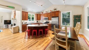 A modern kitchen with wooden cabinets, a central island with stools, a dining table with chairs, and a TV in the corner. - Union Pier Hidden Gem | 3 Blocks to Beach | Screened Porch & Fire Pit (Union Pier)