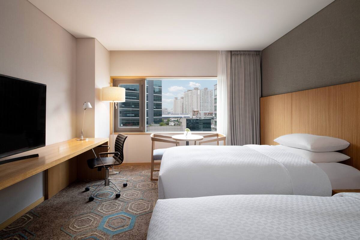 Ein Hotelzimmer mit einem großen Bett, einem Schreibtisch mit Fernseher, einem Sessel, einem kleinen Tisch und einem Fenster mit Blick auf Gebäude.