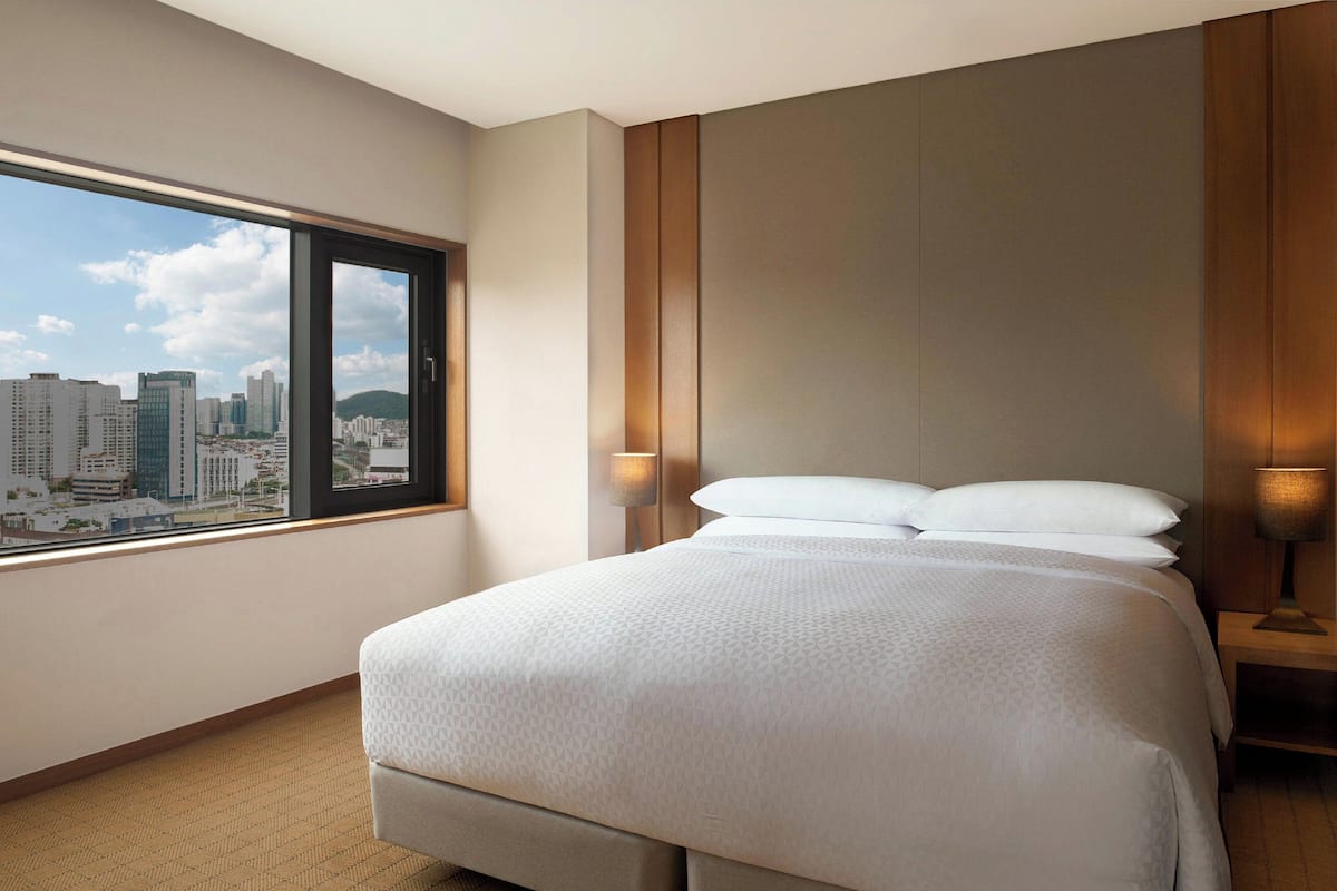 Ein Hotelzimmer mit einem großen Bett, einem Nachttisch, einer Lampe und einem Fenster mit Blick auf die Stadt.