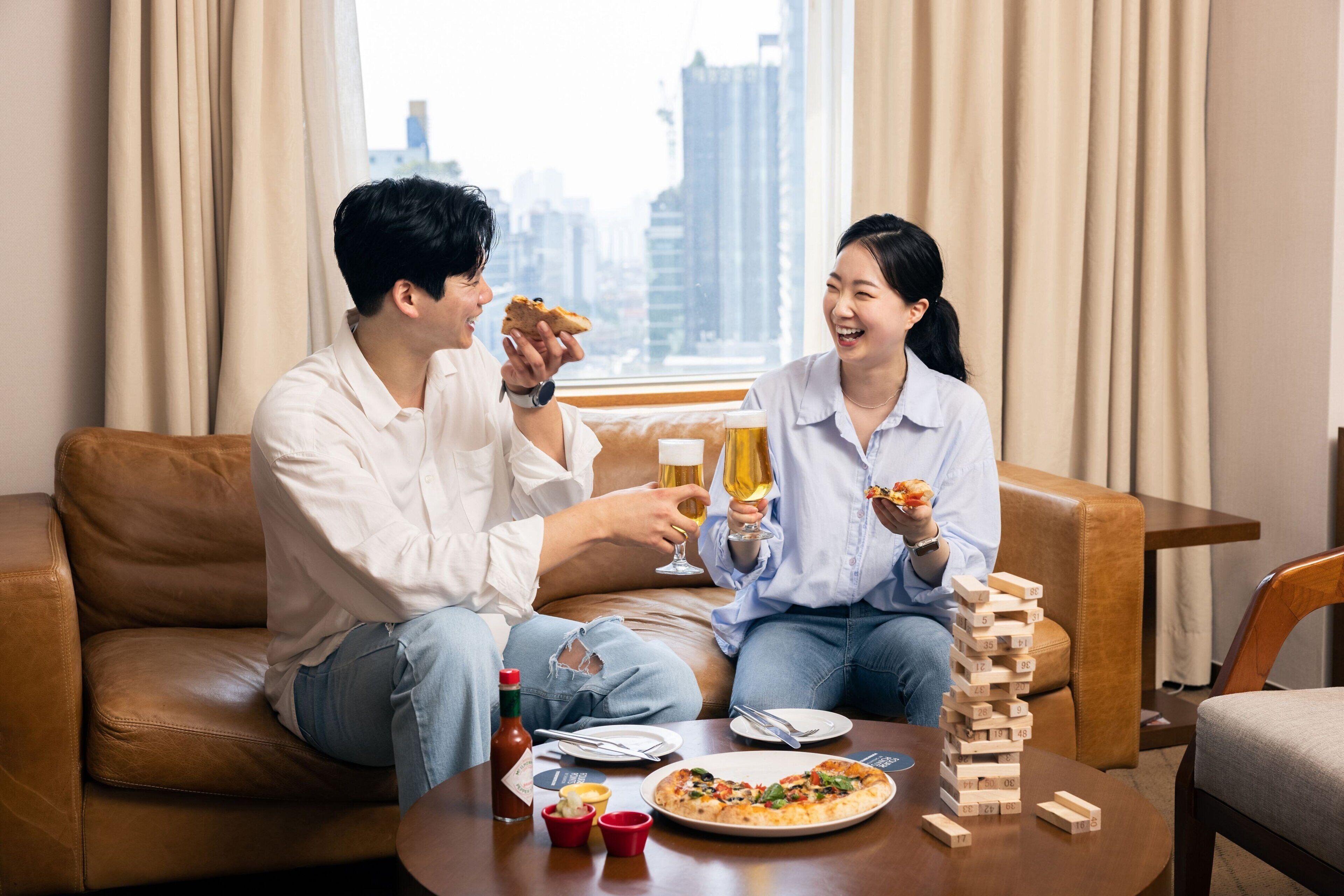 A couple sitting on a brown leather sofa, holding slices of pizza and glasses of beer, with a pizza and a game of Jenga on a table in front of them.