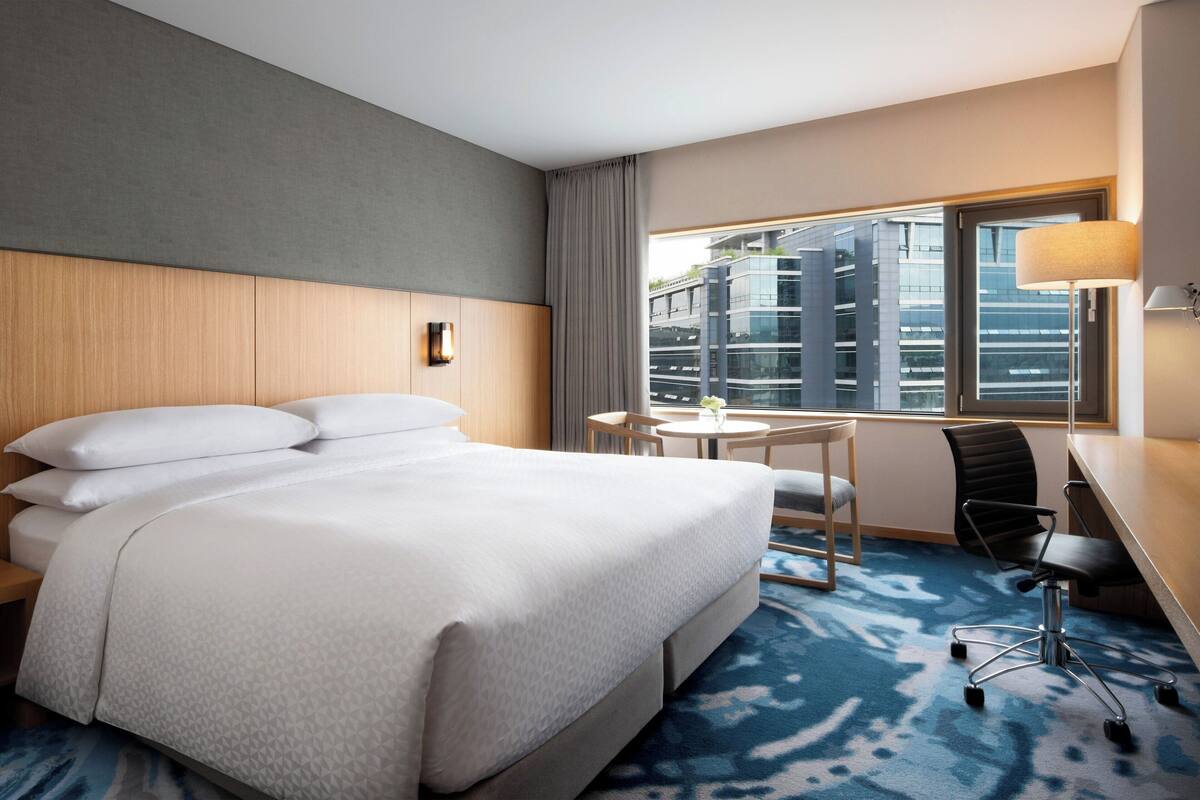 Ein Hotelzimmer mit einem großen Bett, einem Schreibtisch, einem Stuhl und einem Fenster mit Blick auf Gebäude.
