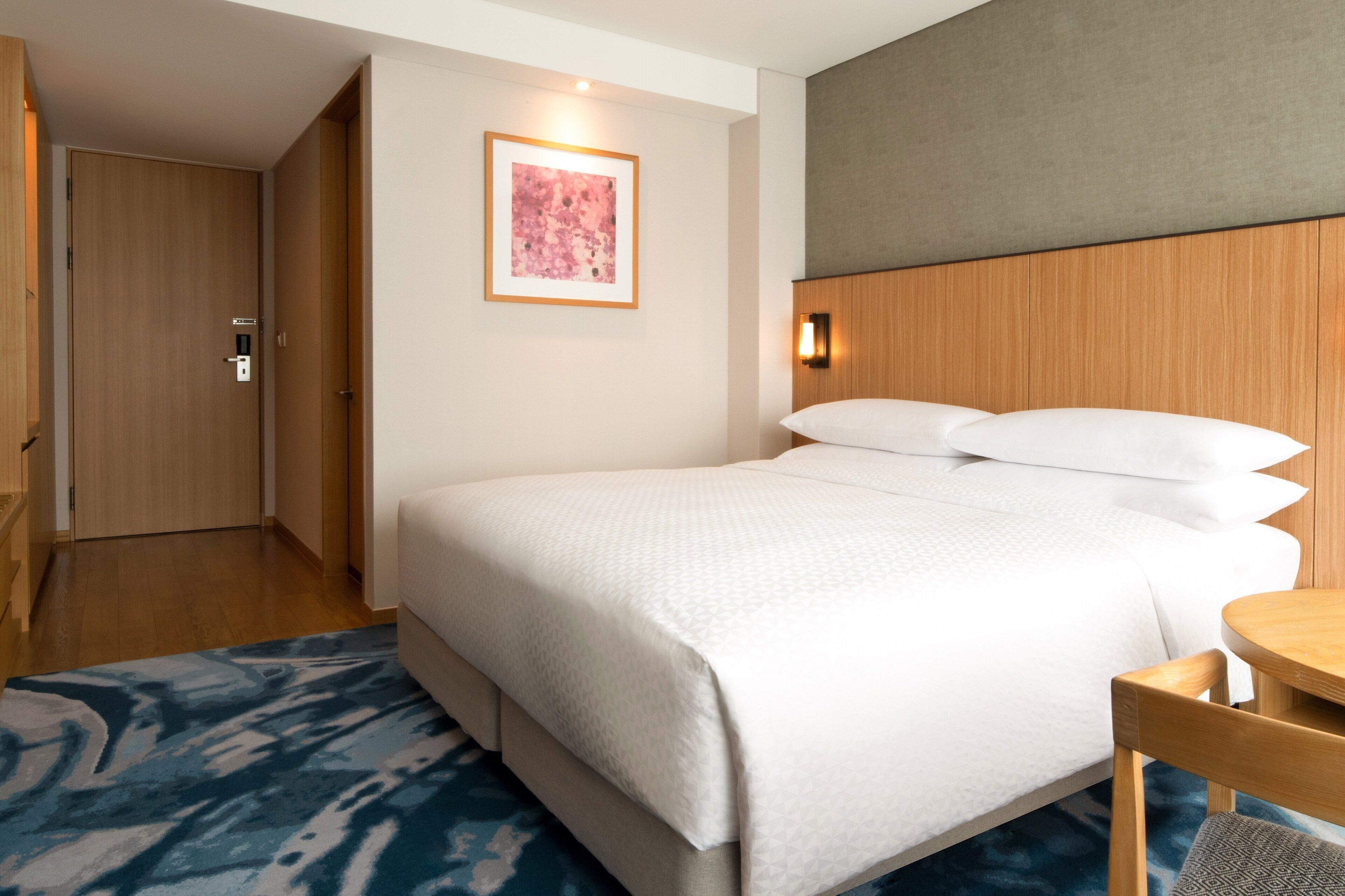 A hotel room with a bed, a bedside table, a chair, a door, and a framed picture on the wall.