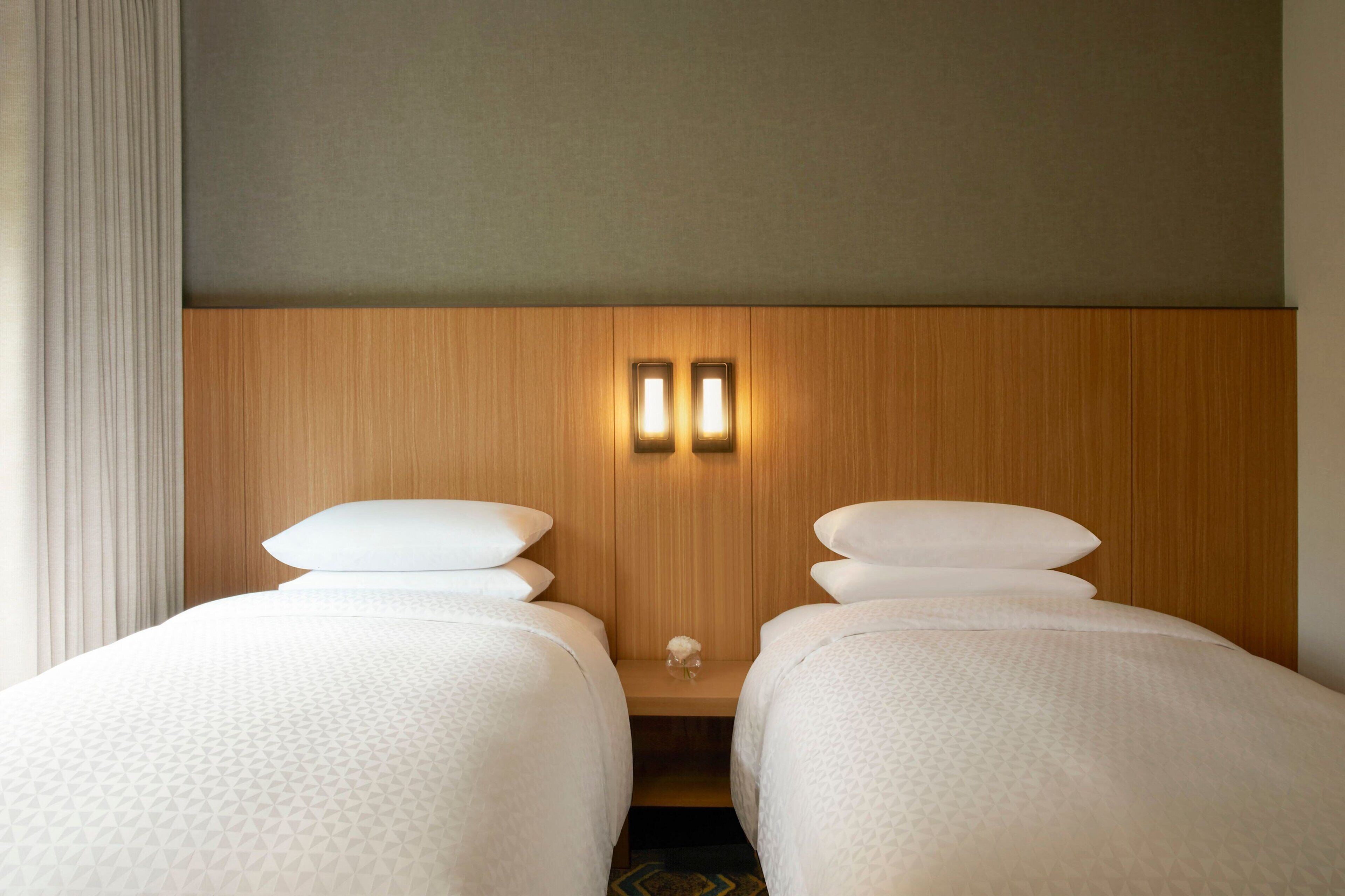 Habitación de hotel con dos camas individuales, cabecero de madera y una mesita de noche con dos luces de pared.