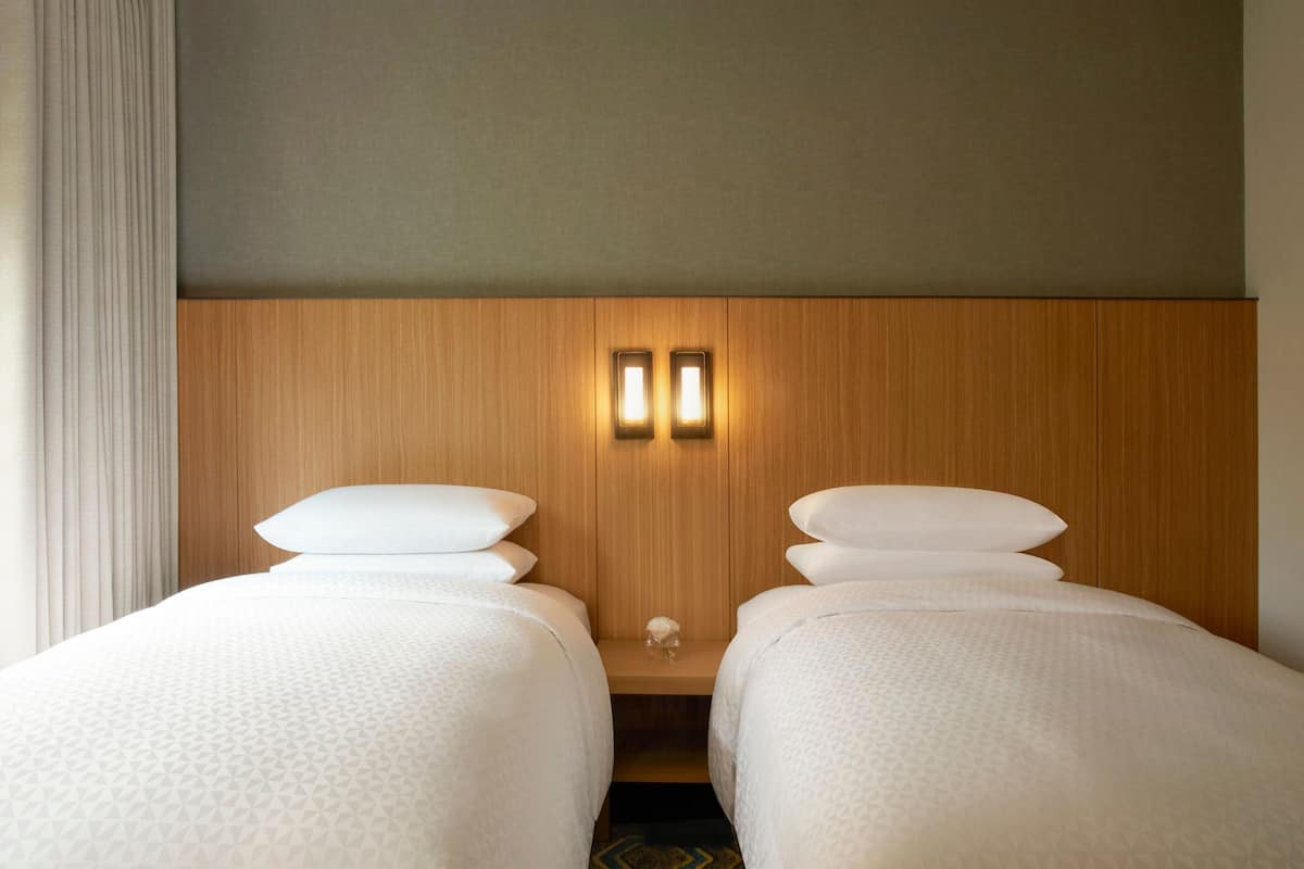 Une chambre d’hôtel avec deux lits simples, une tête de lit en bois et une petite table avec deux lampes fixées au mur.