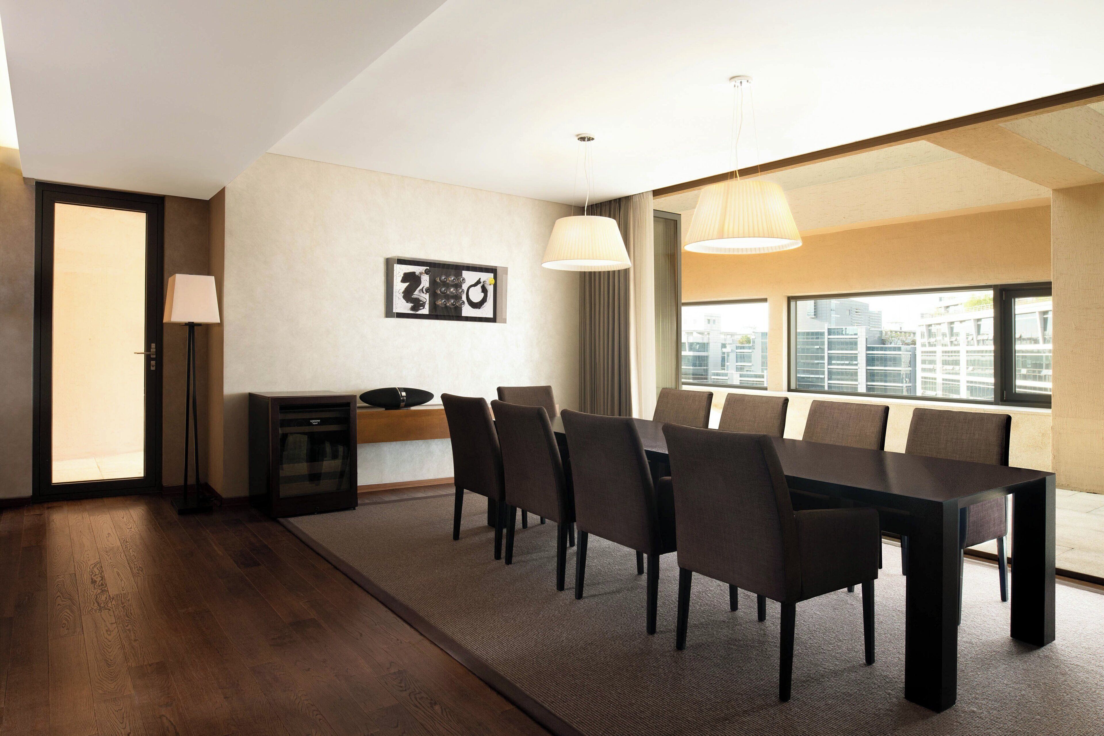 A modern hotel room with a large dining table, comfortable chairs, a lamp, and a view of the city through large windows.