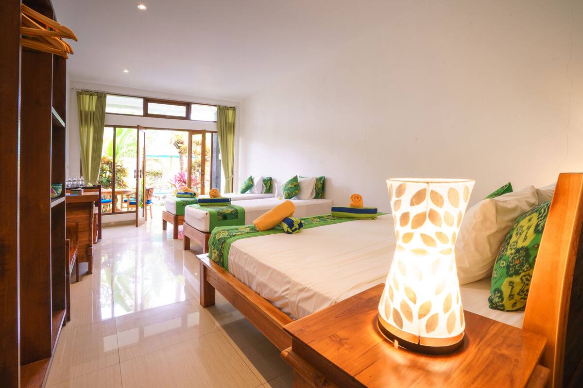 Quarto de hotel com uma cama grande, uma mesa de cabeceira de madeira, uma escrivaninha com cadeira e vista para o exterior através de uma porta de correr de vidro.
