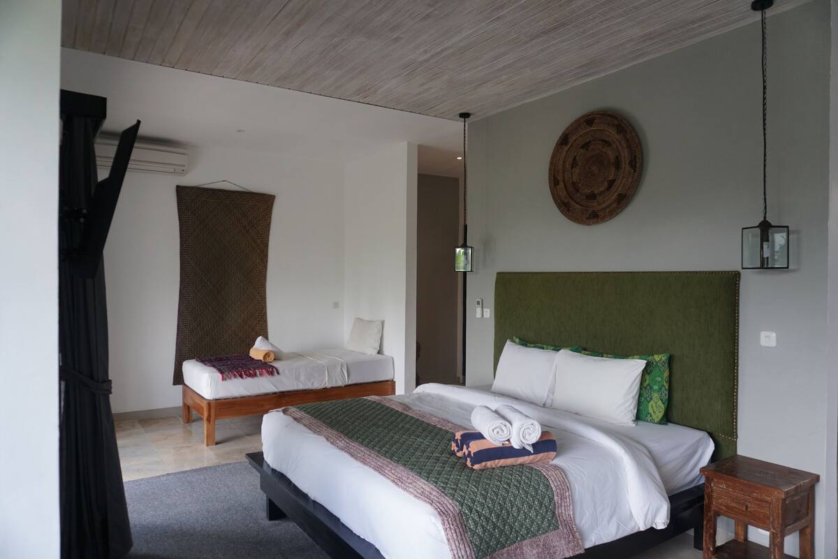 Quarto de hotel com uma cama grande, uma cama menor, cabeceira de madeira, um painel de parede tecido, luminária pendente e uma pequena mesa de cabeceira de madeira.