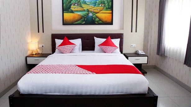Een keurig opgemaakt bed met een rood-witte sprei, twee nachtkastjes met lampen, een schilderij van een landschap aan de muur, en een raam met gordijnen.