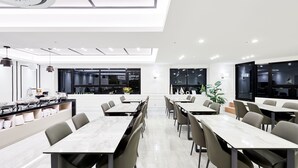 A modern dining area with long tables and chairs, a kitchen in the background, and a view of the city through large windows. - Seattle B Hotel (Busan)