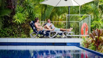 A couple relaxing by a poolside lounge chair with an umbrella, surrounded by lush tropical plants.