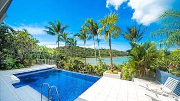 A swimming pool with clear blue water, surrounded by lush greenery and palm trees, overlooking a scenic beach and ocean.