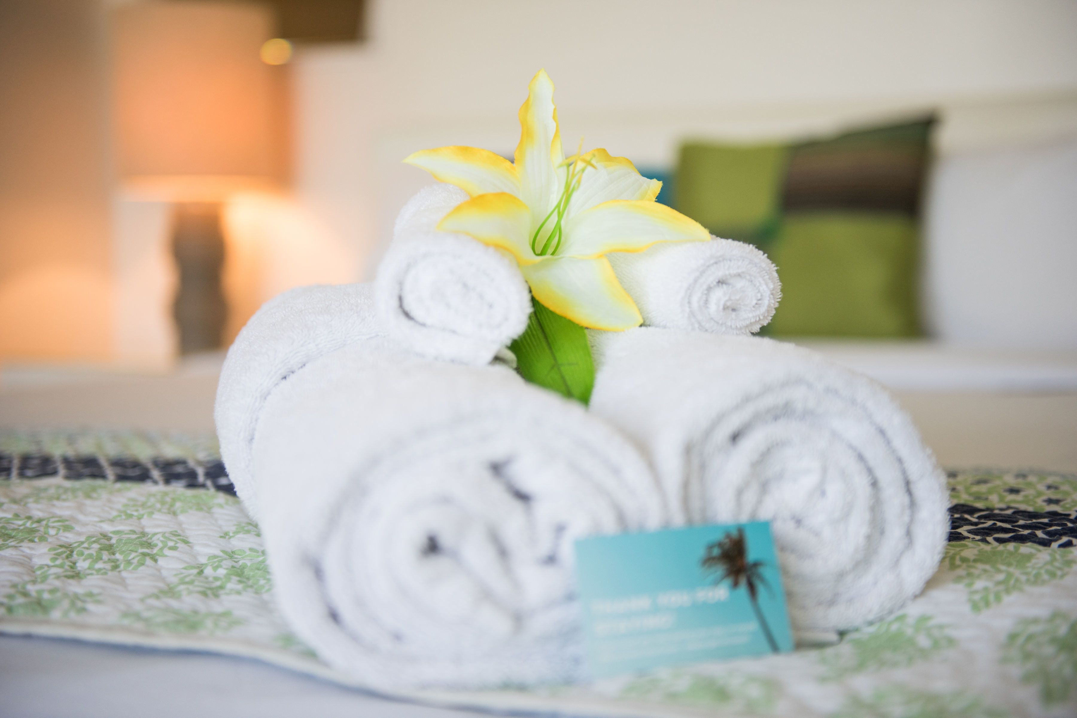 A neatly arranged set of rolled white towels and a single yellow and white flower on a bed with a patterned bedspread.