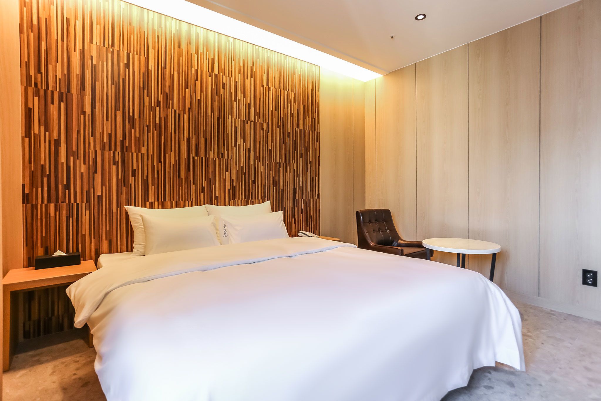 A modern hotel room with a large bed, a wooden accent wall, a small round table, and a chair.