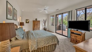 A bedroom with a bed, a television, a ceiling fan, a wooden cabinet, a vase, and a window with a view of trees. - Santa Susana | West Sedona Townhome w/ Pool, Hot Tub & Red Rock Views (Sedona)