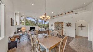 A spacious living area with a dining table, comfortable seating, a fireplace, and large windows offering a view of the outdoors. - Santa Susana | West Sedona Townhome w/ Pool, Hot Tub & Red Rock Views (Sedona)