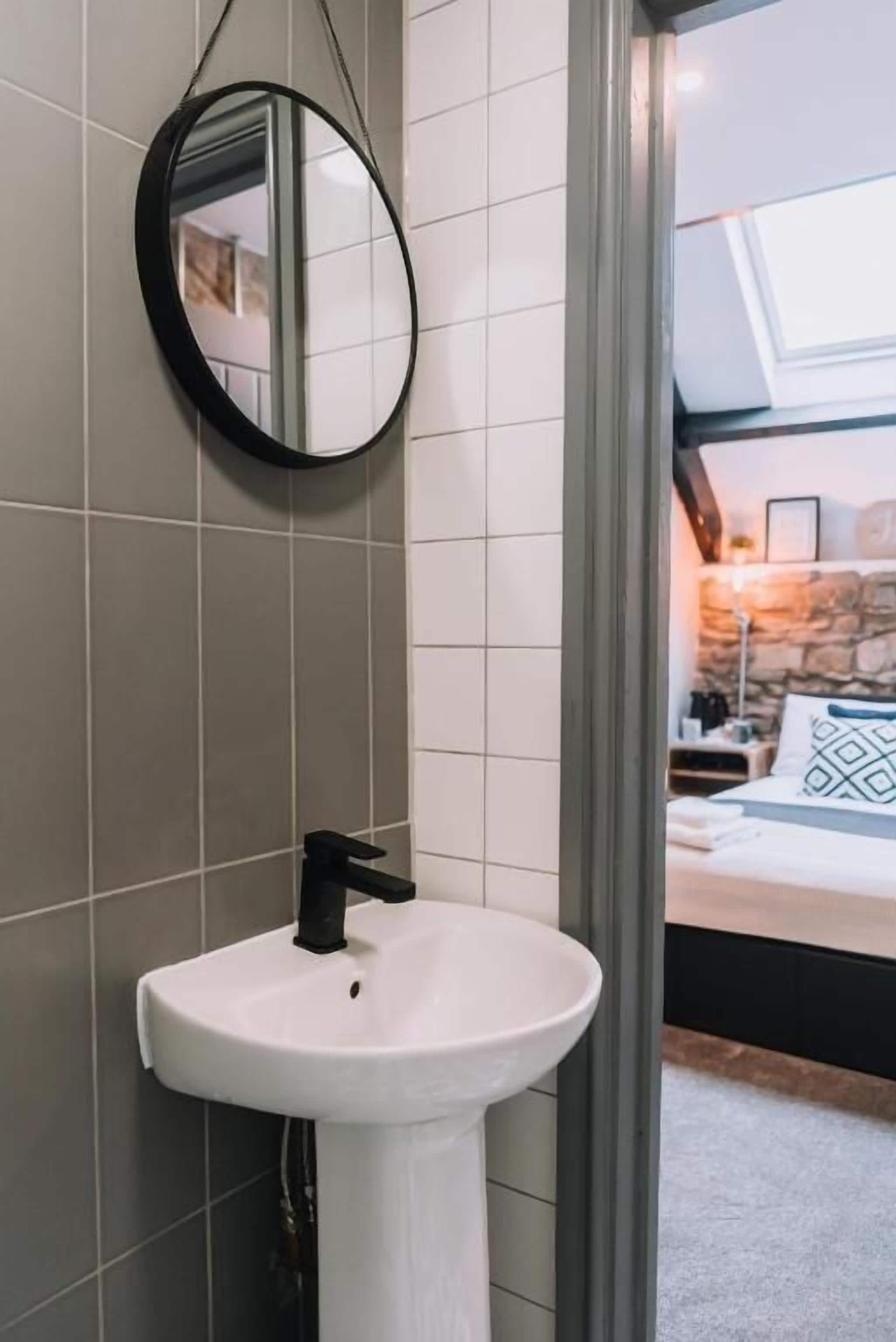 Un baño con un lavamanos blanco, un espejo redondo y vistas a un dormitorio con cama y pared de piedra.