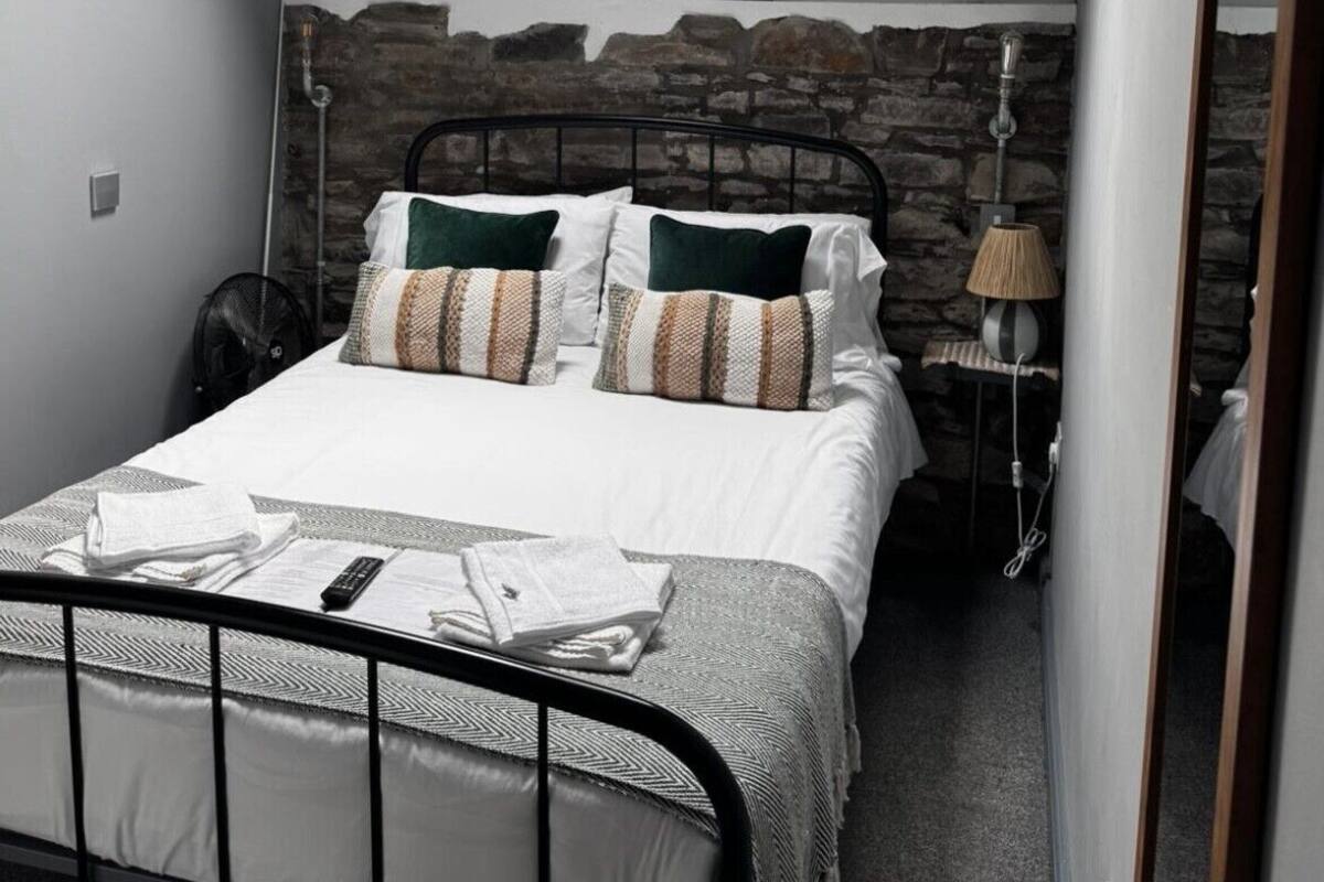 Una cama individual con estructura metálica, ropa de cama blanca y almohadas estampadas. Se ve una lámpara pequeña y un calentador de pared.