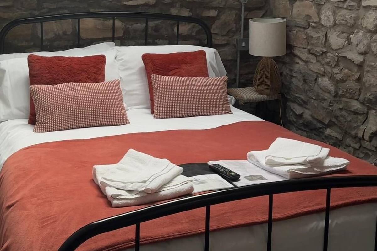 Un dormitorio con cama, almohadas, una lámpara de noche, un cartel que dice 'Be our guest' y una pared de piedra.
