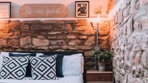 A bedroom with a stone wall, a bed with patterned pillows, a bedside table with a lamp, and a 'Be our guest' sign above the bed. - Sgwd Gwladys (Neath)