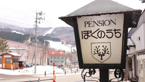 A pension sign with a tree emblem, a snowy landscape, and buildings in the background. - Zao Onsen Pension Bokunouchi (Kaminoyama)