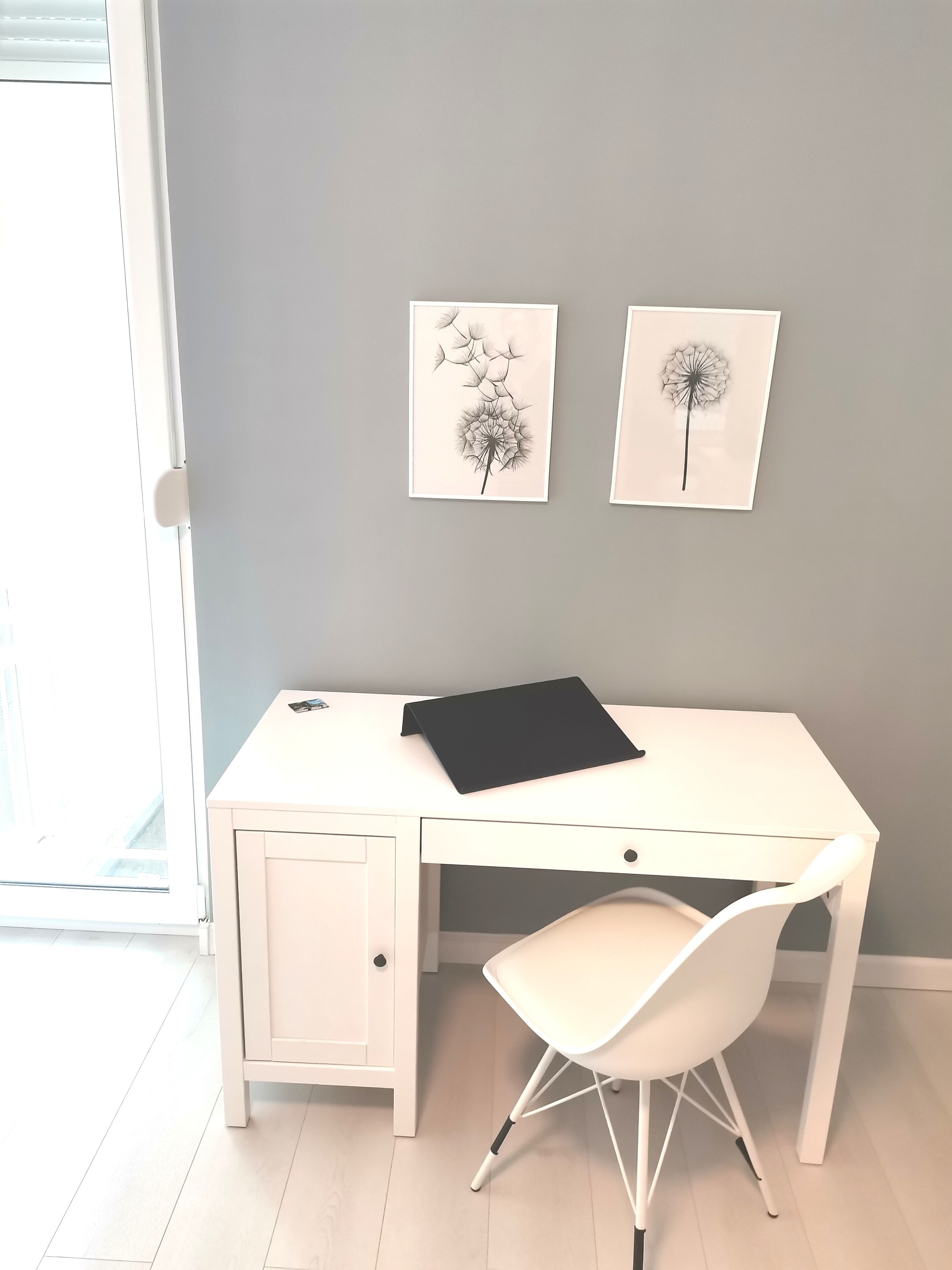 Ein weißer Schreibtisch mit einer schwarzen Ordnermappe, ein weißer Stuhl und zwei gerahmte Bilder von Löwenzahn an einer grauen Wand.