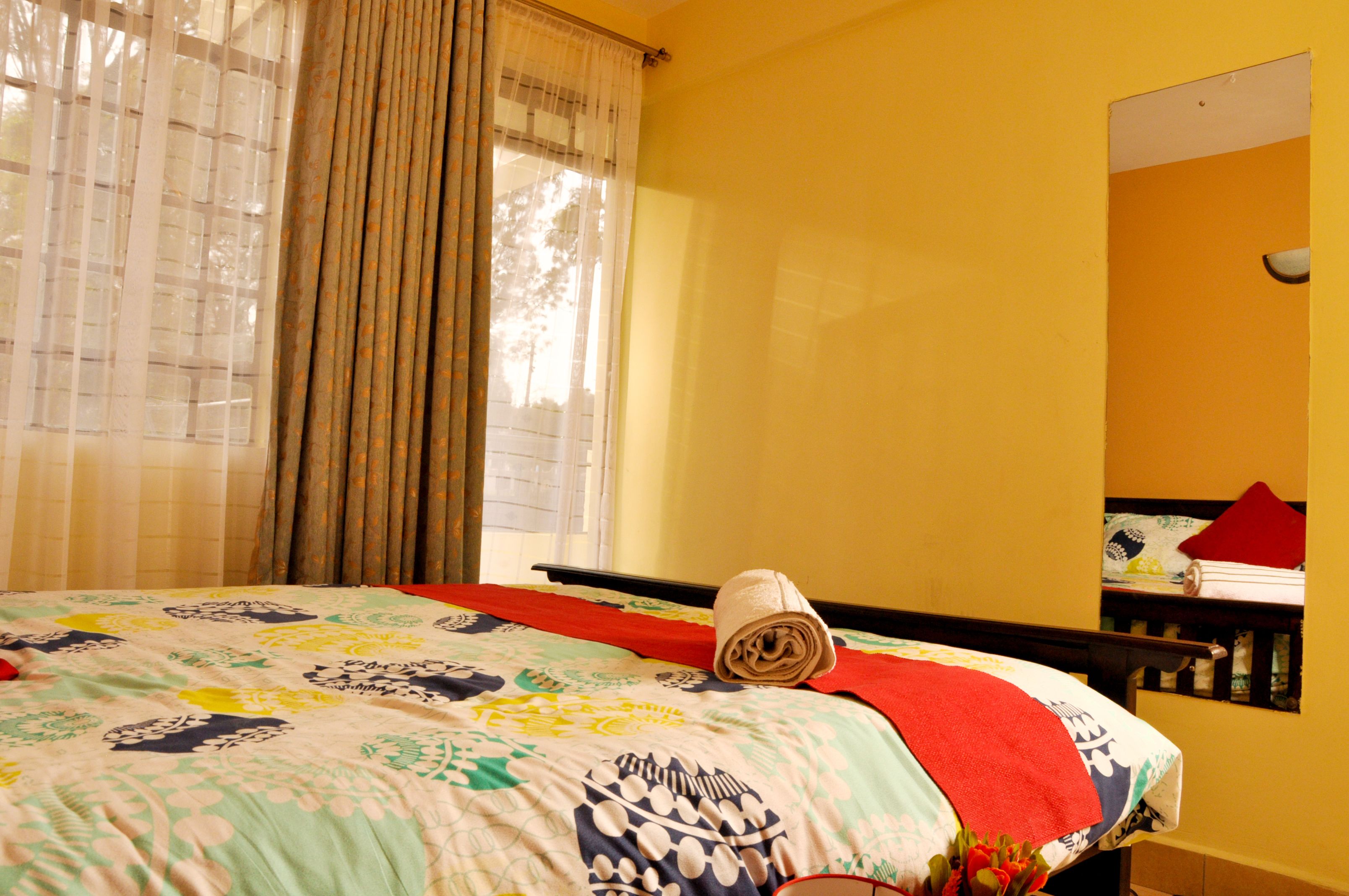 A hotel room with a bed, a window with curtains, a red towel on the bed, and a mirror on the wall.