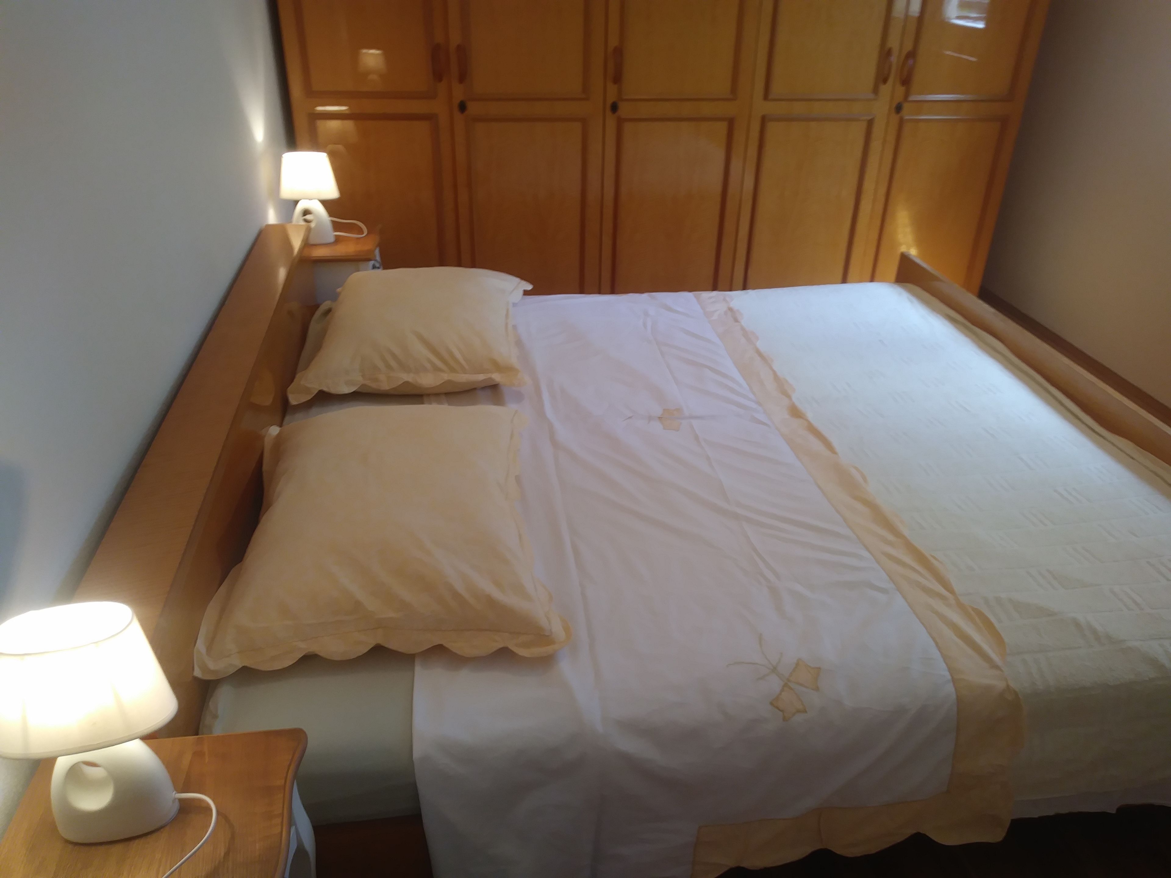 Una cama bien hecha con una colcha blanca y beige, dos almohadas y una lámpara de noche sobre una mesita de madera.