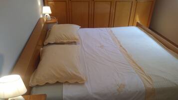 Una cama bien hecha con una colcha blanca y beige, dos almohadas y una lámpara de noche sobre una mesita de madera.
