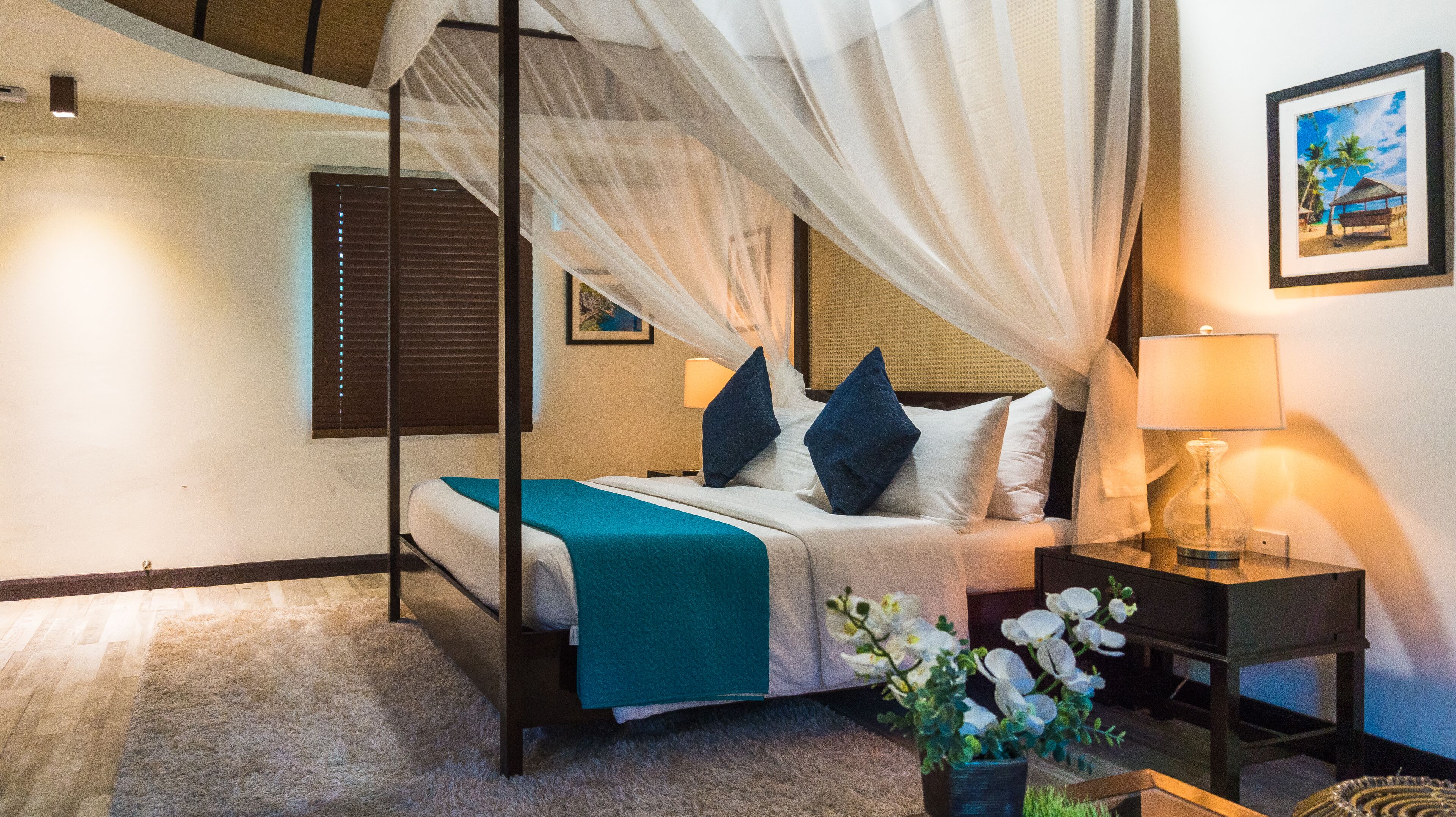 A bedroom with a four-poster bed, a nightstand with a lamp, a framed picture on the wall, and a window with blinds.