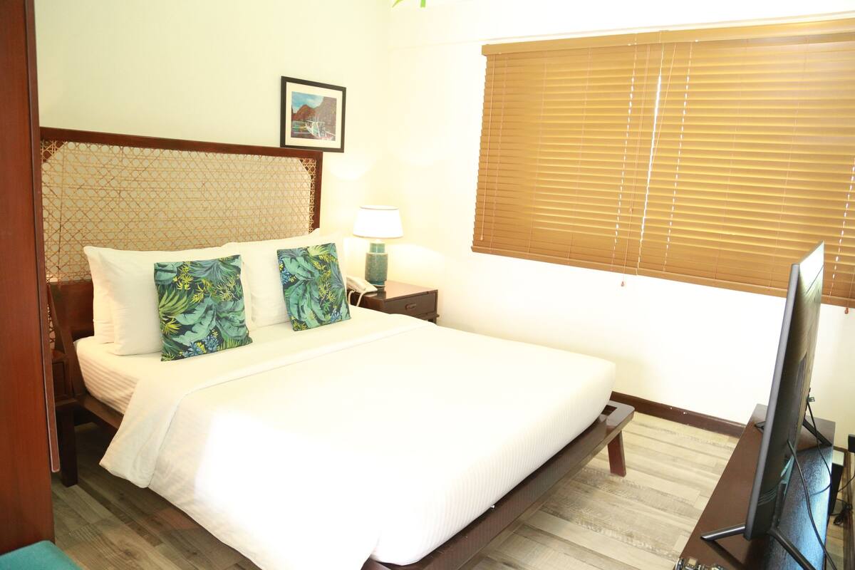 A neatly made bed with white bedding and two decorative pillows, a bedside table with a lamp, a television on a stand, and a window with blinds.