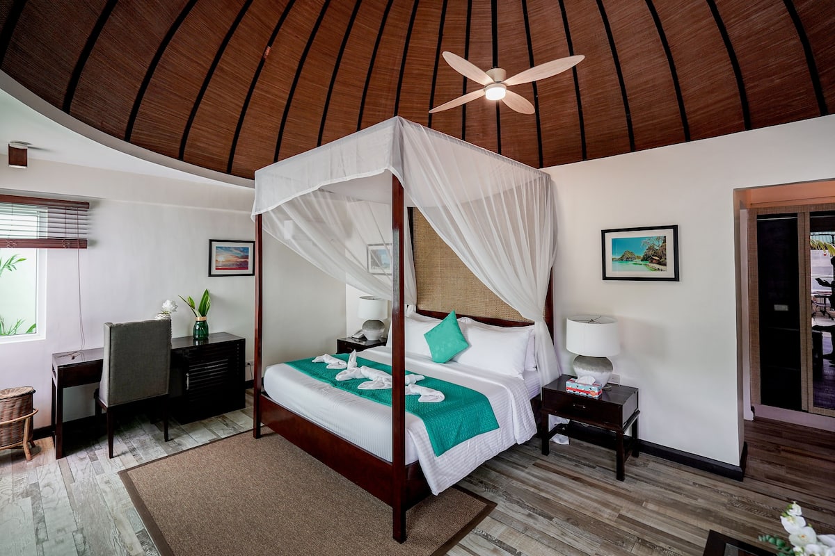 A four-poster bed with a canopy, a desk with a chair, a nightstand, a wall-mounted TV, and a window with blinds.