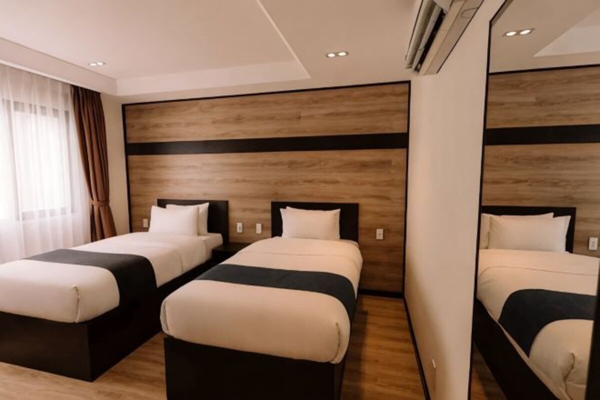 Une chambre d’hôtel avec deux lits, une tête de lit en bois, un grand miroir et une fenêtre avec des rideaux.