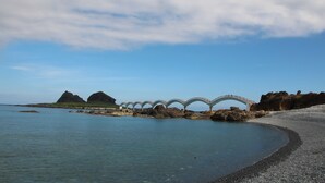 A coastal scene with a white bridge over water, rocky outcrops, and a pebbly beach.