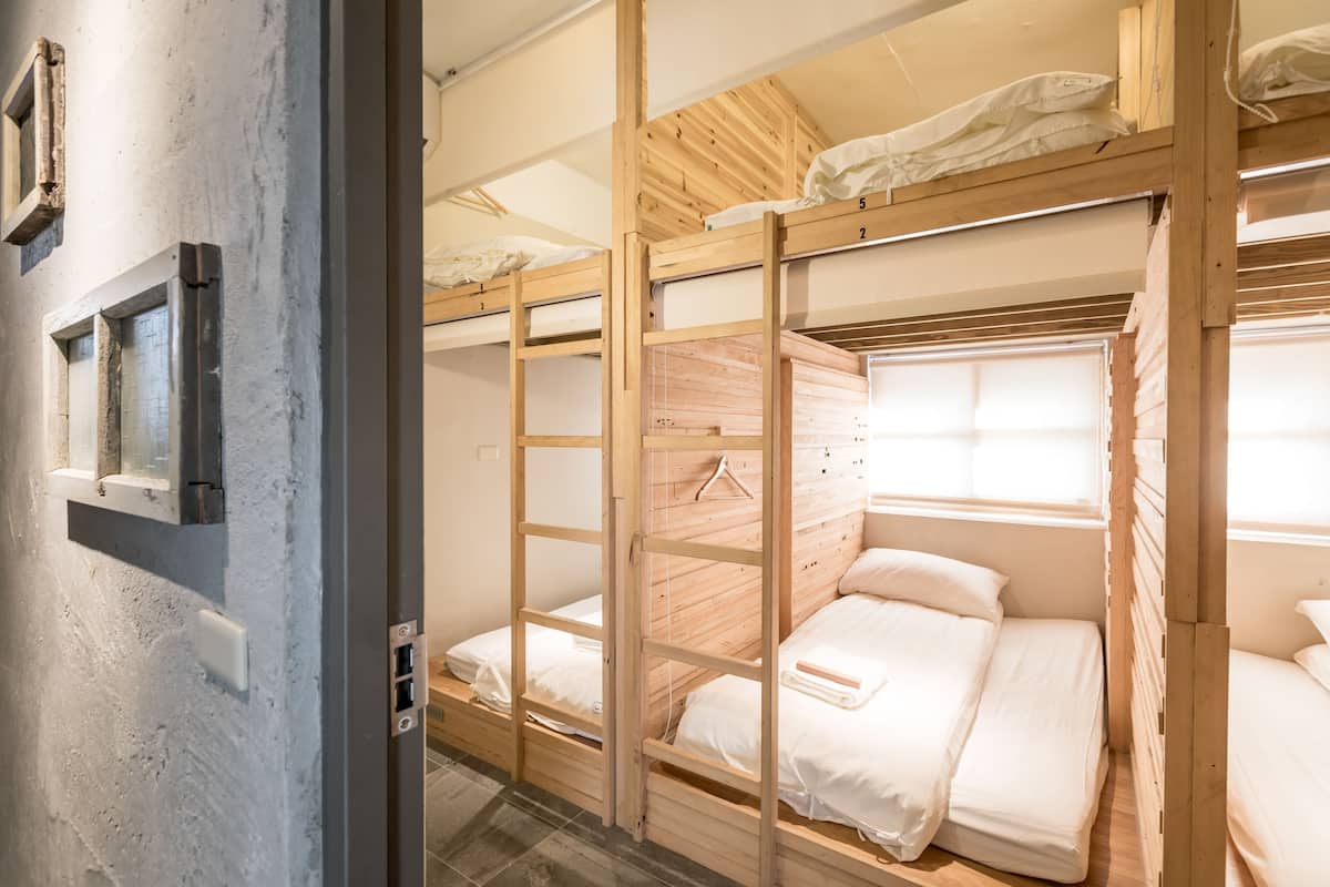 A bunk bed room with wooden frames, white bedding, and a window with blinds.