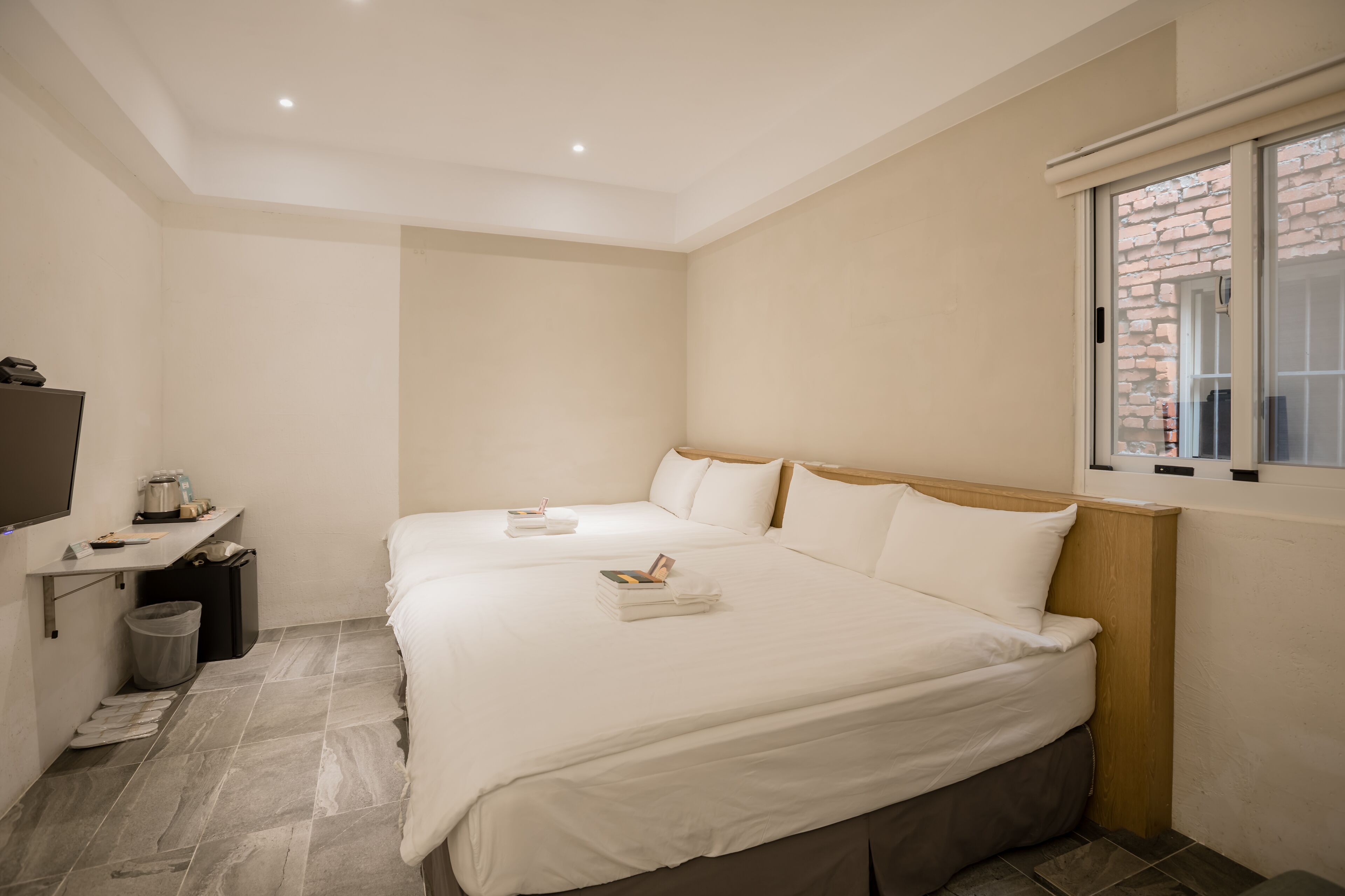 Habitación de hotel con dos camas, un escritorio pequeño con una silla, un cesto de basura y una ventana con vistas a un muro de ladrillos.