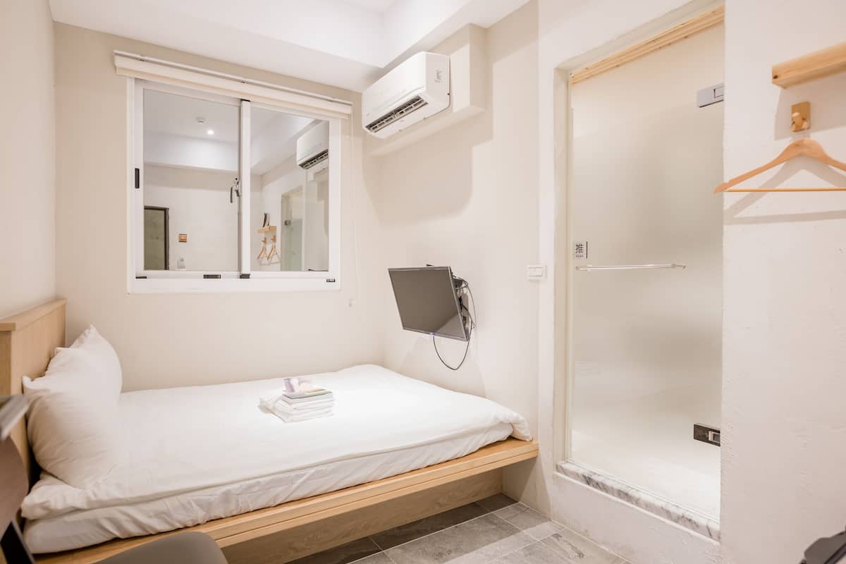 A hotel room with a bed, a bedside table, a mirror, an air conditioning unit, a shower, and a wooden hanger.