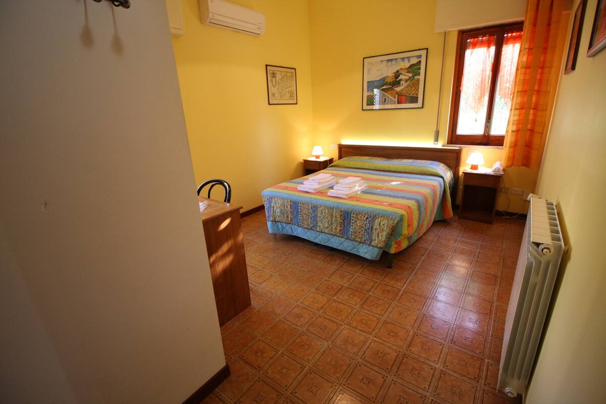 A hotel room with a bed, two bedside tables, a chair, a desk, and a radiator.