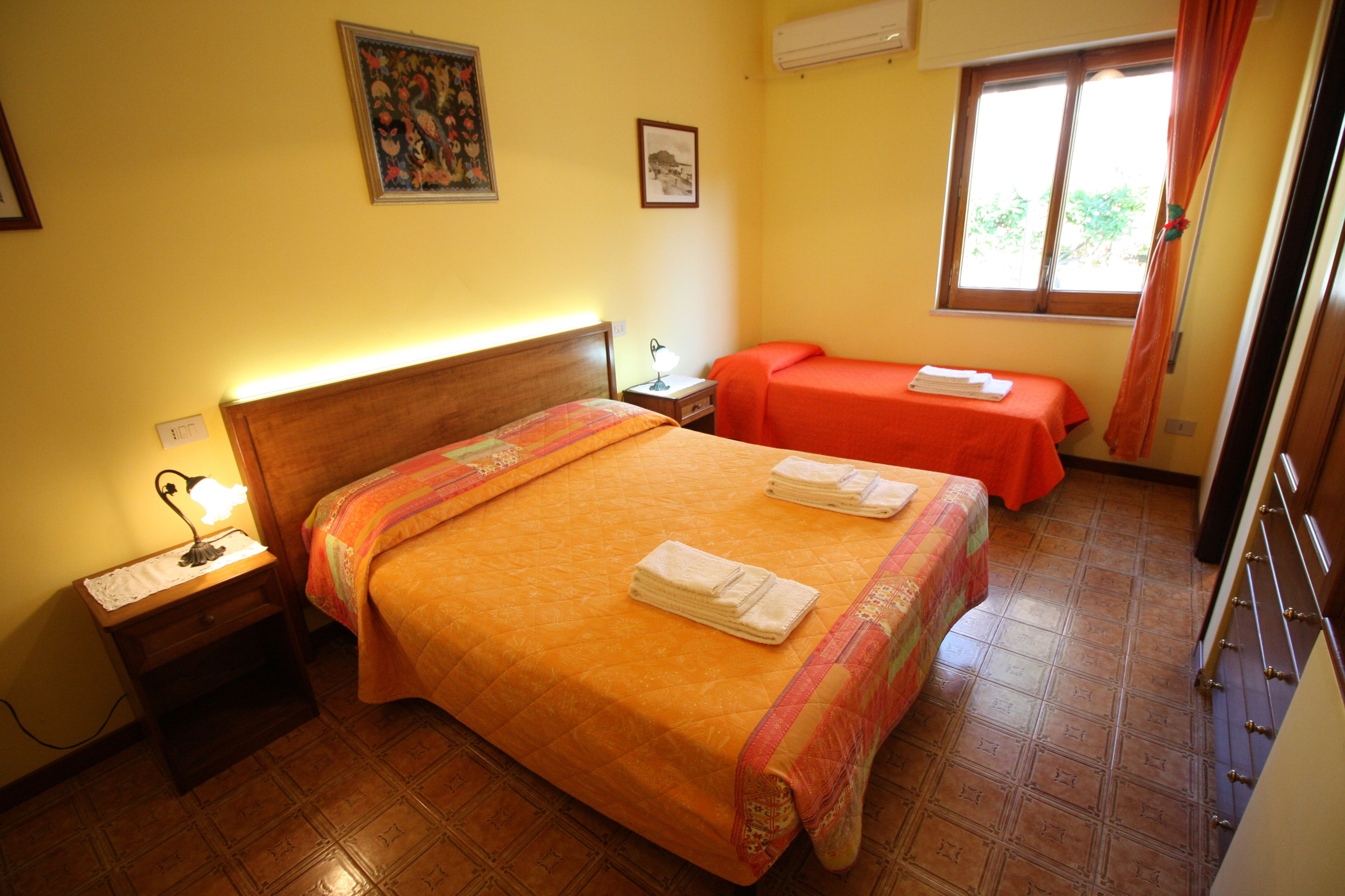 Una camera d'albergo con un letto grande, un letto più piccolo, un comodino con una lampada, una finestra con tende e una parete con due quadri incorniciati.