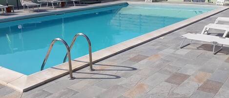 A swimming pool with a tiled deck, lounge chairs, and a wooden fence in the background.