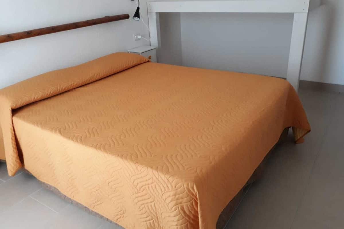 Un lit recouvert d’une couvre-lit orange, dans une chambre dont la tête de lit est blanche et où l’on aperçoit une poutre en bois au mur.