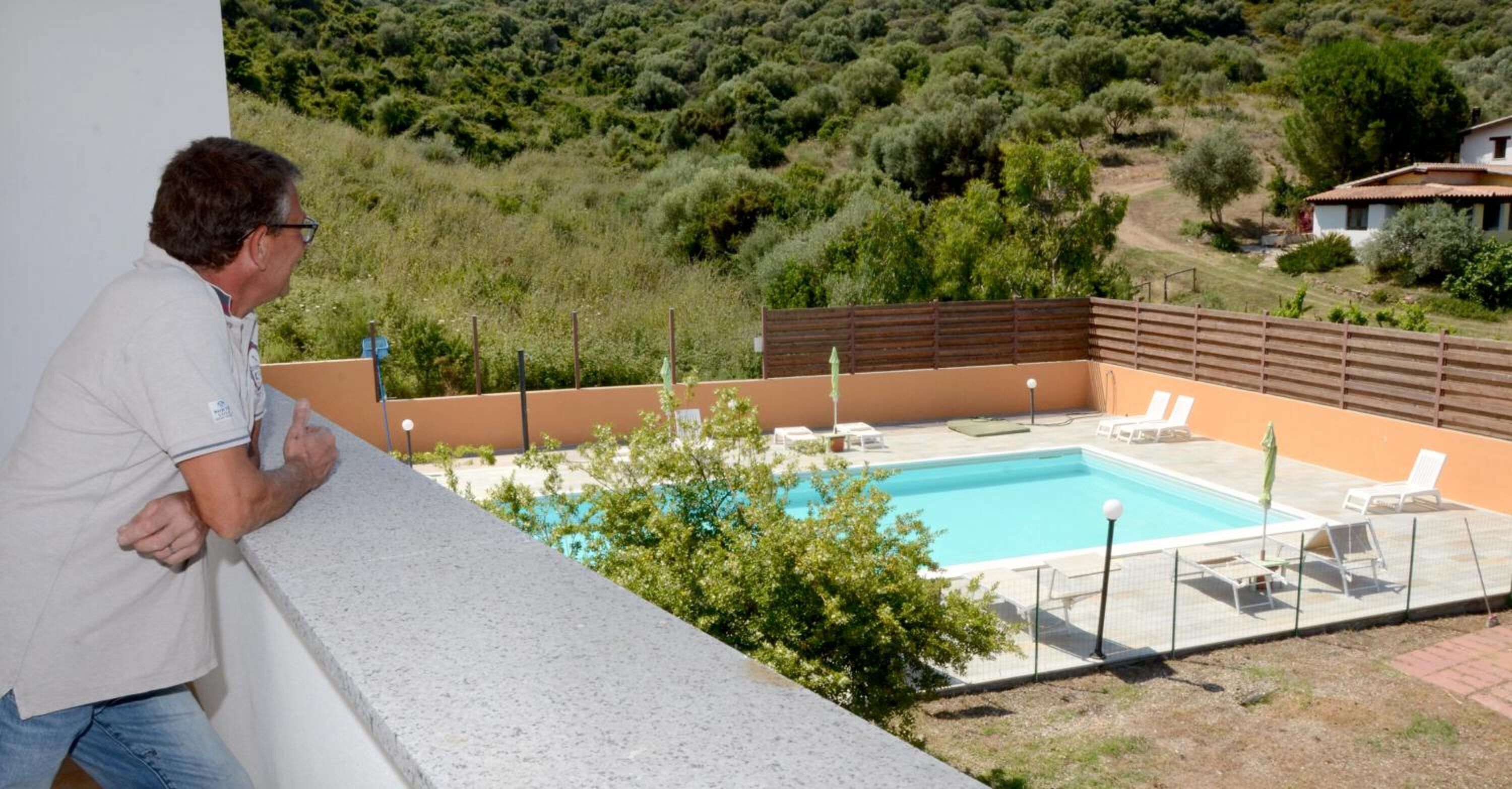 A man overlooking a swimming pool with lounge chairs and a wooden fence.