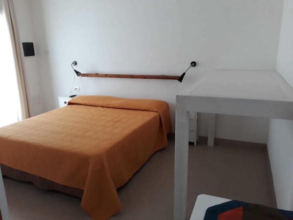 Une pièce simple et épurée, avec un lit, un bureau blanc et une étagère fixée au mur.