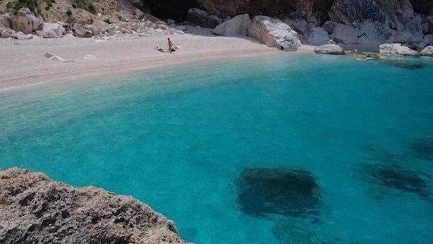 Een kuststrand met helder turquoise water, rotsachtig terrein en een persoon die over het zand loopt.