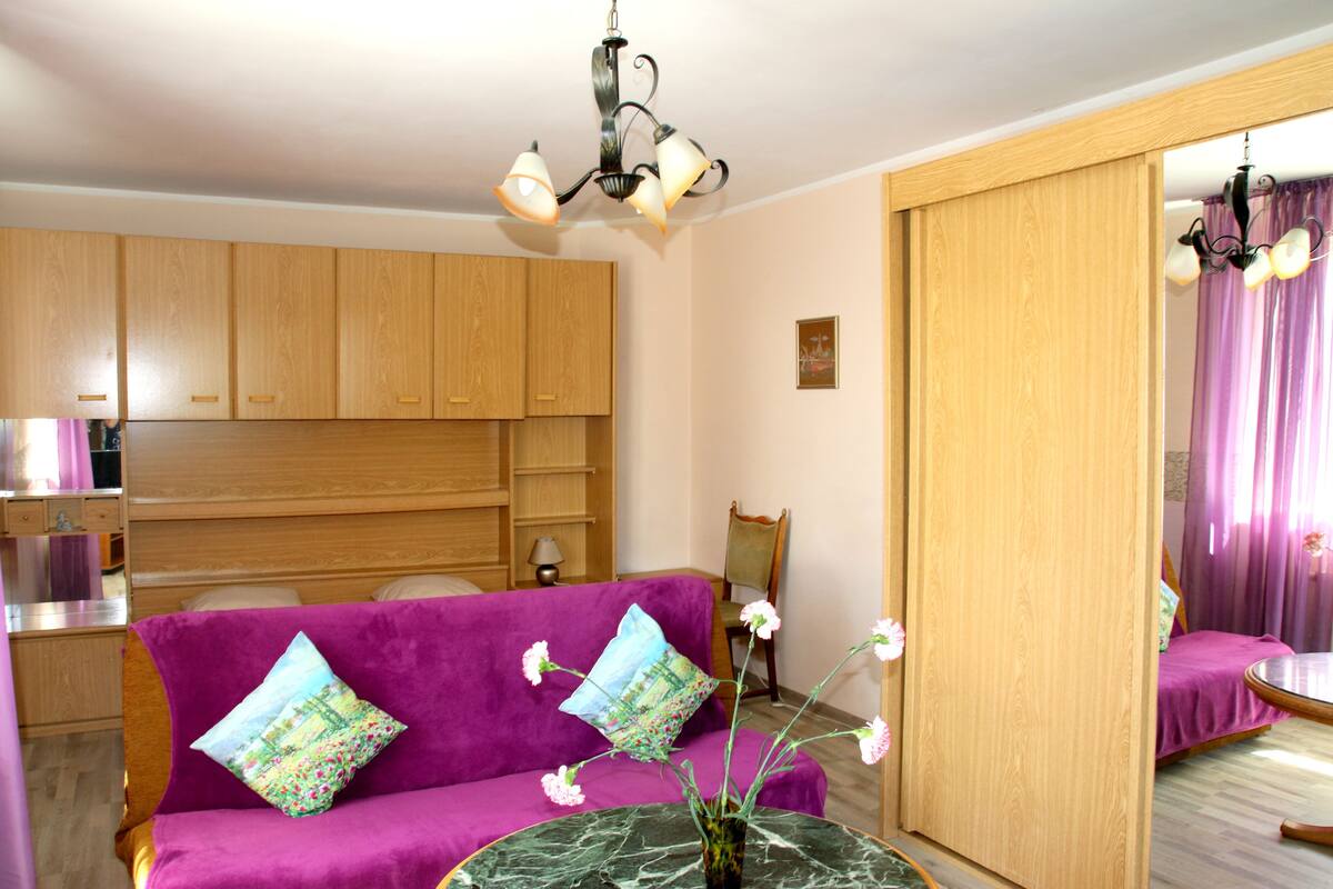A room with a purple sofa, a small table with a vase, wooden cabinets, a chair, and a mirror.