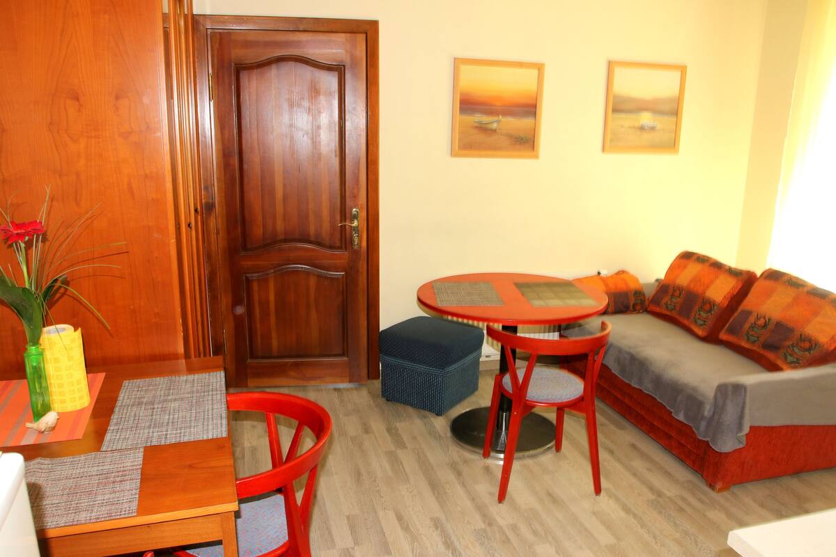 A room with a wooden door, a round table with two chairs, a sofa with cushions, and two framed pictures on the wall.