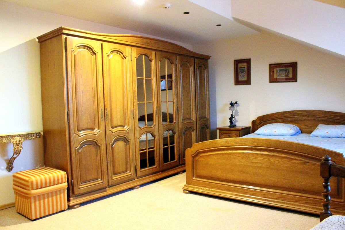 A bedroom with a large wooden wardrobe, a bed with a wooden headboard, and a small ottoman.
