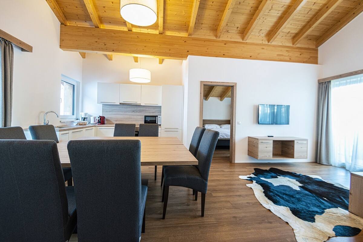Een moderne woonkamer met een houten plafond, een eethoek met een grote tafel en stoelen, een keuken op de achtergrond en een televisie aan de muur.