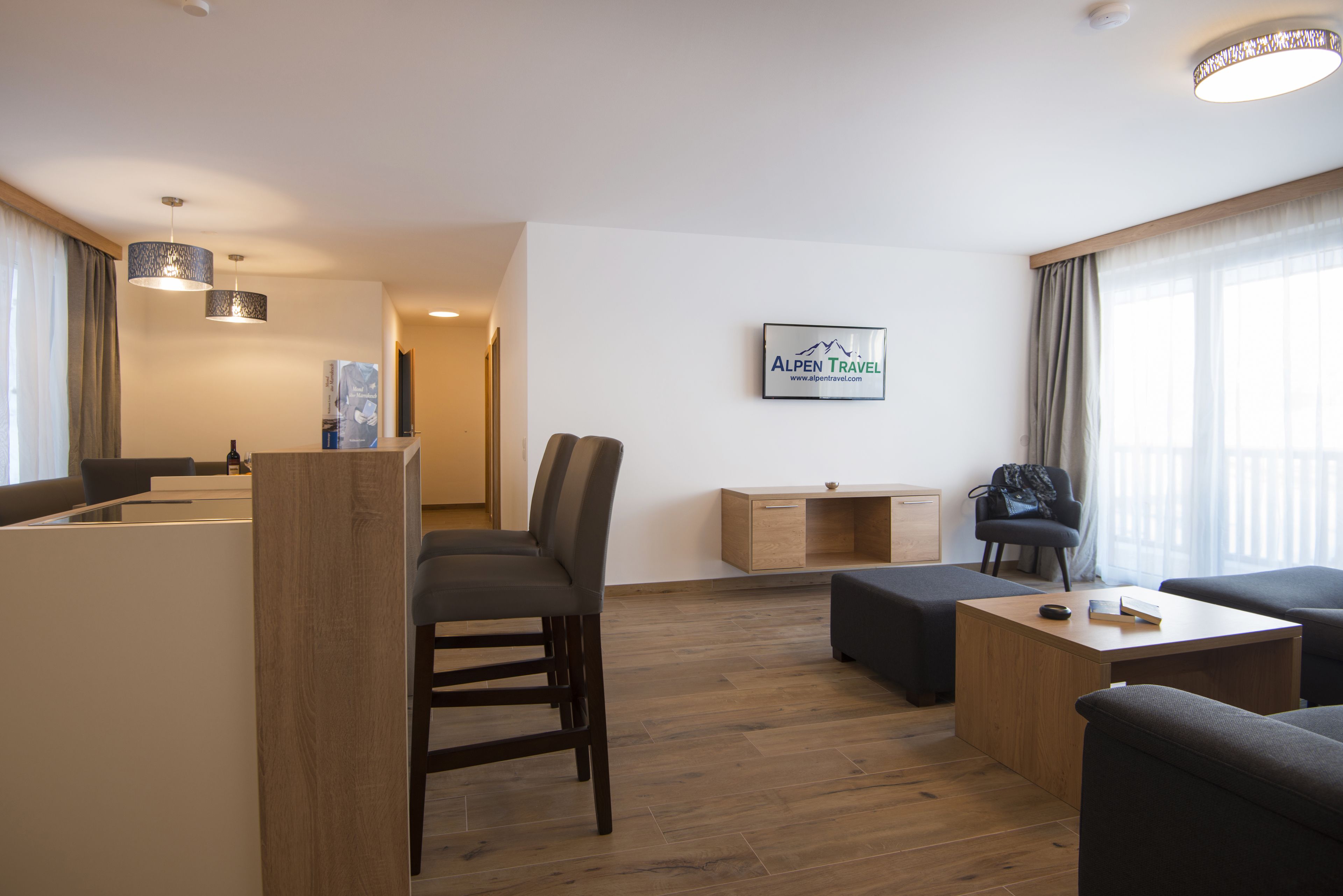 Quarto de hotel moderno com piso de madeira, área de refeições com mesa e cadeiras, um sofá, uma pequena mesa lateral de madeira e uma placa fixada na parede.
